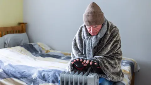 An older man wearing a woolley hat, scarf and blanket indoors huddled over an electric fire in a bedroom 
