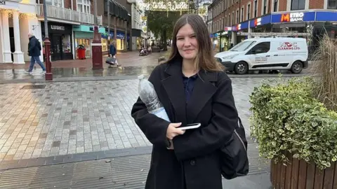 Phil Harrison/BBC A woman with long dark hair and a dark overcoat stands in the middle of Tunbridge Wells holding a large plastice bottle of water.