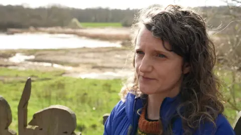 Caucasian woman with mousy blonde and brown curly hair. She has blue eyes and is wearing an orange jumper and blue raincoat. She is standing in front of the nature reserve's marshes where you can see a lake in the background and green grass.
