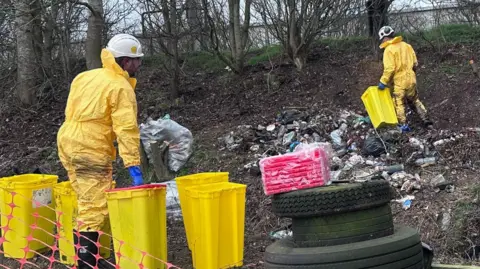 People in protective suits work on a clearup of waste on a woodland verge close to a road. 