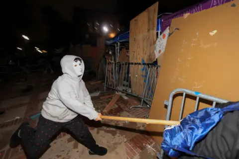 A man in hoodie and mask attacks a barricade with a wooden pole