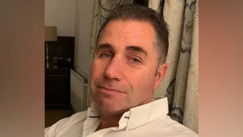 A middle-aged man with dark short hair that us greying at the sides. He is looking toward the camera and is wearing a white button down shirt. He is sitting in front of some cream curtains with a floral design. 