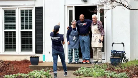 Shawn Harris, wearing a campaign t-shirt with his name on it, poses with a middle-aged couple in their gardening aprons in front of their house while door-knocking. A volunteer, also in a campaign-shirt takes a picture