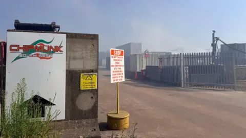 The entrance of Chipmunk South West's site. There is a large white sign at the entrance with the company logo and an arrow pointing forward. There is a stop sign with instructions for vehicles entering the site and a yellow caution sign. Smoke can be seen in the sky from the fire.