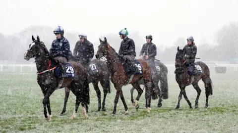 Horses striding in the snow on Cheltenham Racecourse, with jockeys sitting on top