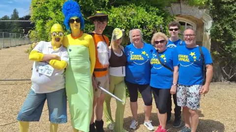 Katy Griffin Someone dressed as Homer Simpson in a yellow bald-cap. She is joined by someone dressed as Marge Simpson with big blue hair and they stand next a woman in an Ogre-mask dressed as Shrek and people in blue t-shirts which say 'Stars Appeal.'