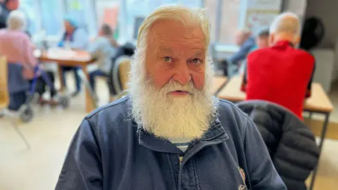 William Anderson, sitting down in a community centre. He is wearing a blue top and has a white beard.