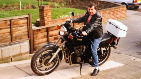 Supplied Robert is wearing a black leather jacket and jeans and is sitting on a Yamaha motorbike on a driveway to a house.