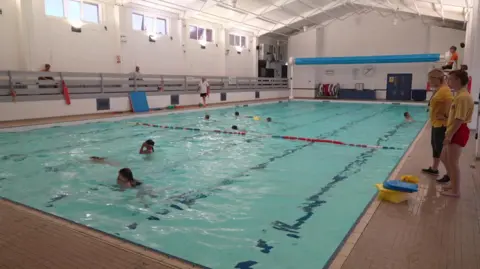An indoor pool with two instructors at the side and several children swimming.