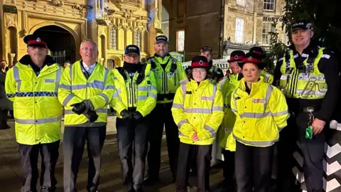 GRASAC A group of police officers, including Gloucestershire Police and Crime Commissioner Chris Nelson, stand wrapped in hi-vis coats and warm uniform while standing outside a church in Cirencester's Market Place, next to a Christmas tree.