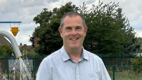 Ian Taylor stares at the camera. He wears a light blue shirt. In the background is a large green tree and metal fencing. To the left are parts of a splash pad. 