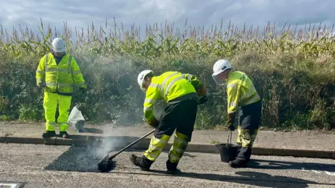 Workmen in hi-vis spreading material over a section of road to fix potholes.