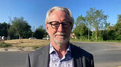 Martin Johnson poses for a photo. He is wearing a black suit jacket over a patterned shirt, with dark glasses. He has a neatly trimmed white beard and mid-length white hair. A roundabout can be seen in the background.