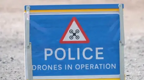 A fold-up portable sign in blue sits on the ground. It features a red warning triangle with text saying: "Police drones in operation".