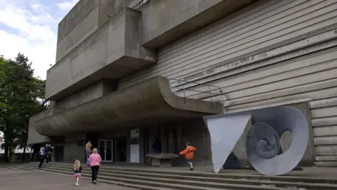 A large gray building with pacemaker blocks and horizontal straps is a swirly gray metal sculpture above stages. Some people are going inside.