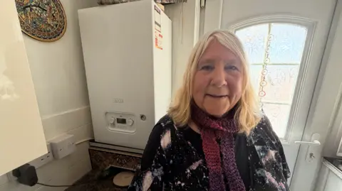 Lynda Graves, who is wearing a black star-patterned shirt and a red and purple knitted scarf, stands in front of her new white boiler. It has been installed in her kitchen after the previous boiler became faulty leaving her without any heating or hot water.