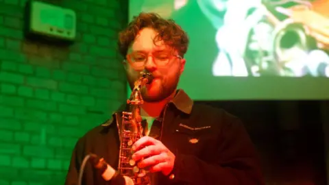 John Bray James has brown curly hair and is wearing orange glasses. He is wearing a black coat and is playing the saxophone. 
