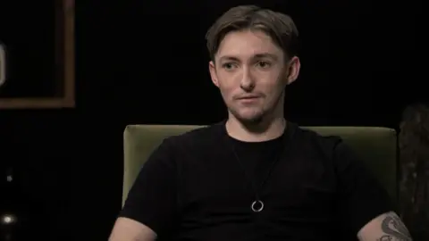Matthew Johnson is wearing a black T-shirt and is sitting in a chair looking towards an interviewer out of shot.