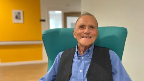 A man with short grey hair is smiling at the camera. He is wearing a grey wool waistcoat over a blue shirt. He is sitting in a turquoise chair.