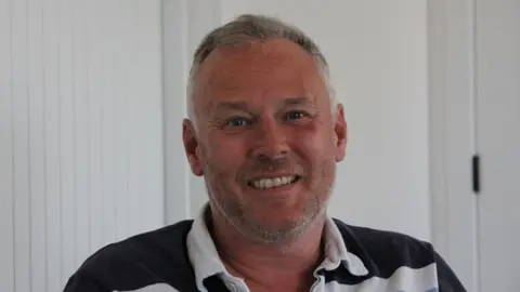 A man with short grey hair and grey stubble looks at the camera smiling. he is wearing a white and black polo neck top. In the background is a white door and wall.