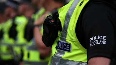 PA Media Row of Police Scotland officers, all unidentifiable. It is focused on the Police Scotland embroidery on the officer's black t-shirt. The officers are wearing hi-viz police vests and have radios. 