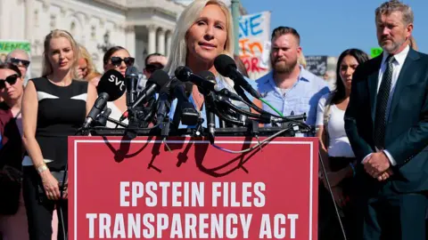 Marjorie Taylor Greene stands at a podium emblazoned with the words 'Epstein Files Transparency Act'. People are stood behind her. 