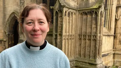 The Rev Becky Lumley wearing a light blue jumper over a vicar's black shirt and white dog-collar stands in front of a corner of a medieval stone church. She has brown hair, tied back, and is smiling.