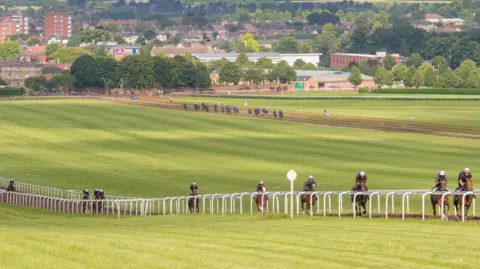 The Newmarket Thoroughbred Racing and Breeding Industry Forum A row of horse riders riding behind one another on a racecourse. 