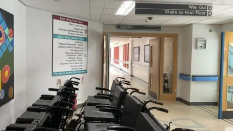 Two rows of black wheelchairs lined up in the corridor of Noble's Hospital. The walls are white with signs giving directions to wards and double doors in the background lead to another empty corridor.