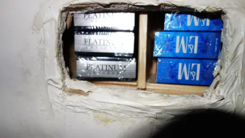 Trading Standards Six cartons of cigarettes are stacked on top of each other in a hidden area of the shop. Three packets of a brand called Platinum and three L&M packets are seen behind a white sliding shelving section.
