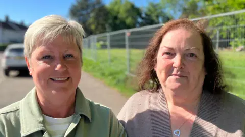 Alana Anderson has short blonde hair and has makeup on. She is wearing a green shirt jacket and a white t-shirt. Dorothy Anderson has long brown hair and is wearing a brown cardigan with a silver necklace. Behind them is a metal railing.