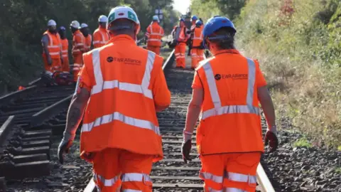 East West Rail Railway engineers make their way along a stretch of track. They are wearing orange fluorescent jackets and trousers