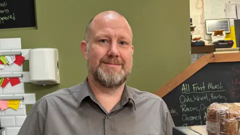 A man with grey hair and a beard, with a grey shirt in a shop with a green wall and some food in plastic wrapping beside him