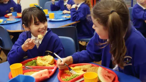 Two school girls are sitting eating their school dinners. One has jacket potato and peas with fruit, and the other has fish and chips with peas and fruit. They are wearing a navy blue school uniform and smiling at each other