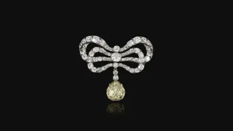 Sotheby's An ornate bow-shaped brooch made of diamonds, with a large yellow diamond drop pendant hanging from the centre, set against a black background.