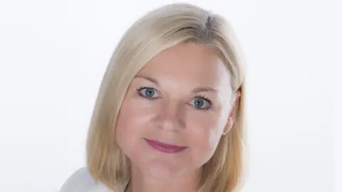 A woman with shoulder-length blonde hair, wearing a white jacket is looking at the camera in a corporate headshot against a white background.