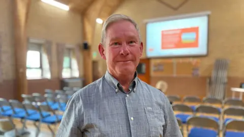 A man wearing a grey shirt is standing in front of rows of chairs inside a building and there is a blurred overhead projector screen in the background.