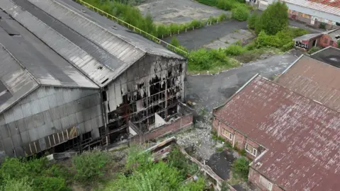 Pictures of abandoned buildings and fire damage. Two buildings that have sustained fire damage are made from corrugated iron and have corrugated iron roofs. It looks like a former factory unit. The front of the buildings have been destroyed by fire. They are in front of a boarded up brick building. The area surrounding it is overgrown with bushes and trees and looks abandoned.