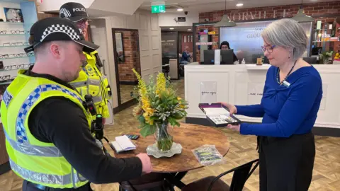 A woman wearing a royal blue top and black trousers is holding out a small box with two sticks of 'SmartWater' inside. She is standing inside an opticians opposite two uniformed police officers.