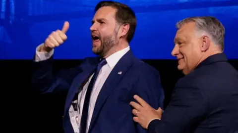 JD Vance and Orban stand together smiling, in front of their national flags