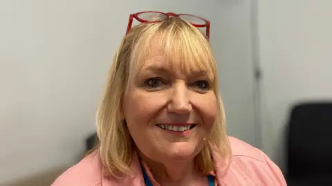 Laura Coffey/BBC A woman with a pink jacket is smiling at the camera. She is wearing a blue lanyard and has blonde shoulder length hair.