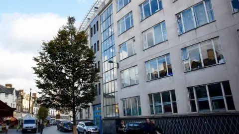Lewisham Council The Old Town Hall is shown as seen from Catford Broadway. It is a 5-storey building which appears quite large. It has a large glass frontage and looks over a residential street where a small market is taking place in the road