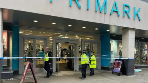 Two police officers and a security guard stand behind police tape at the entrance to a Primark shop 