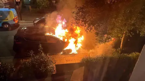 The front half of a car parked on a residential street is seen engulfed in flames. The picture appears to have been taken from inside a neighbours house at night time.