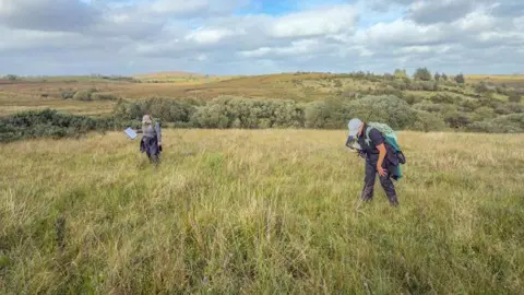 Rose Cremin/Butterfly Conservation Two people dressed in hiking clothing are walking in a large green field. In the distance is green shrubbery and hills. 