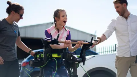 Brook stands, holding onto a horizontal wooden bar. The exoskeleton is black and runs down her legs, with a green bar around her middle. Brooke, whose dark hair is tied in pigtail plaits, smiles, and has a man standing on one side of her and a woman on the other.