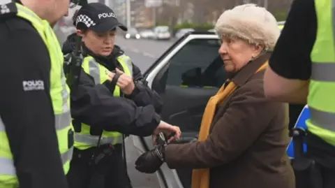 A woman, wearing a brown jacket and cream-coloured puffy hat, is arrested by a female officer next to a police car. Two other officer stand on either side of them in the foreground.