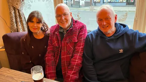 A woman and two men are sitting together looking into the camera smiling. Both men have short white hair, while the woman has long brown hair. Rob in the middle is wearing a red checked shirt and there is a pint of beer on the table.