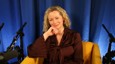 A smiling blonde woman in a brown shirt sits on a yellow armchair, with her head resting on her right arm