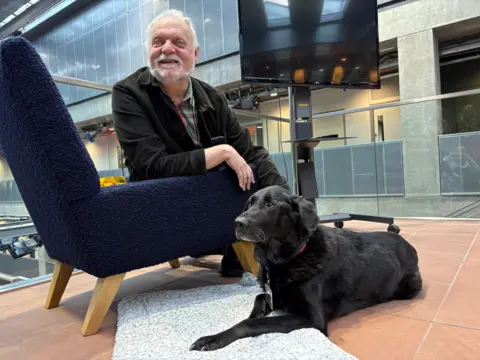 Major the dog is lying in front of Ian Hamilton, who is sitting on a navy blue armchair in the BBC Scotland headquarters at Pacific Quay, Glasgow.